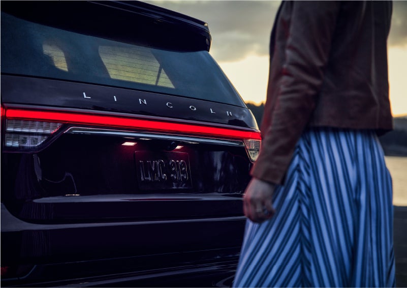 A person is shown near the rear of a 2023 Lincoln Aviator® SUV as the Lincoln Embrace illuminates the rear lights | Brinson Lincoln of Corsicana in Corsicana TX